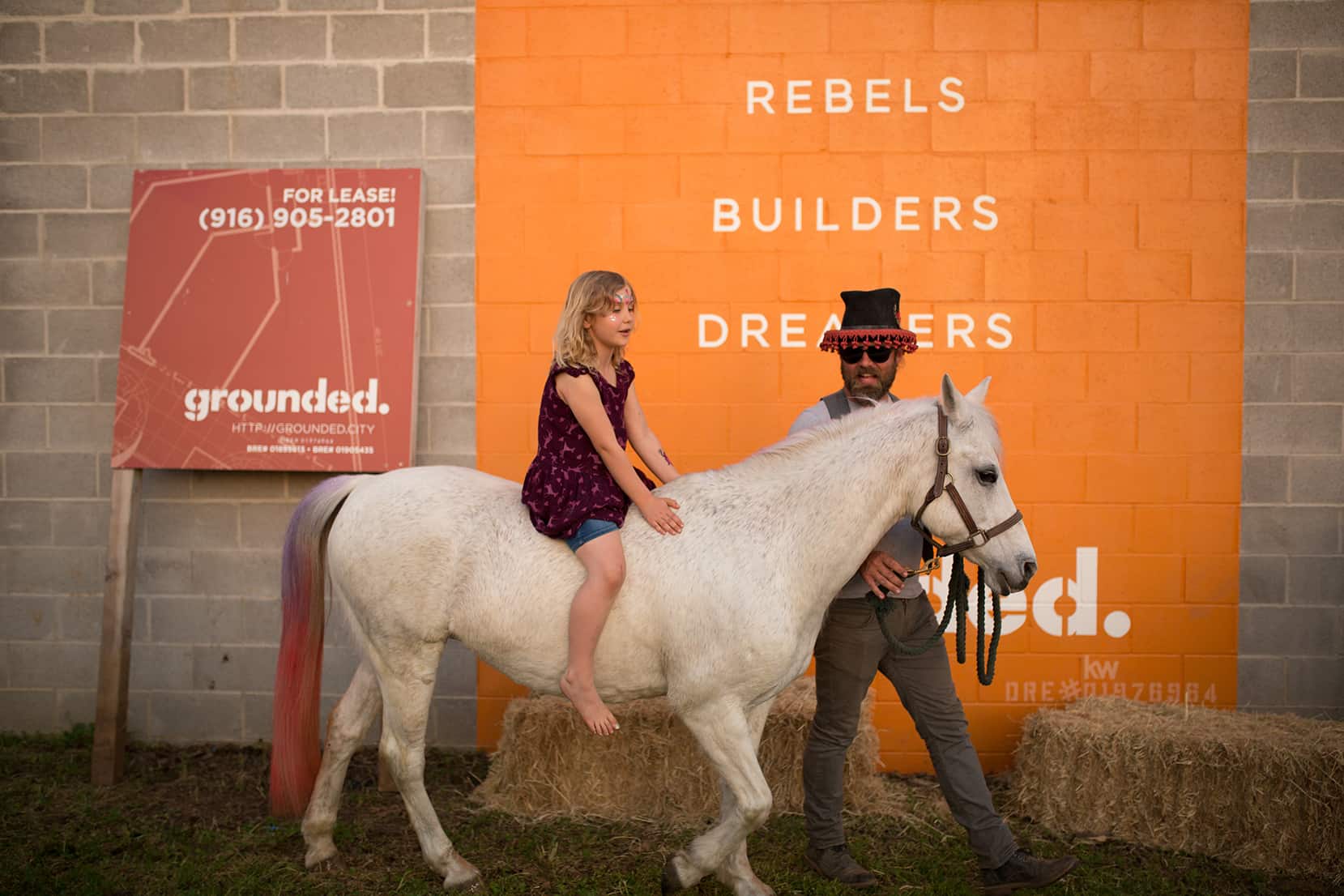 A white horse with a young child riding it, a man in a top hat, and the orange Grounded mural in the background